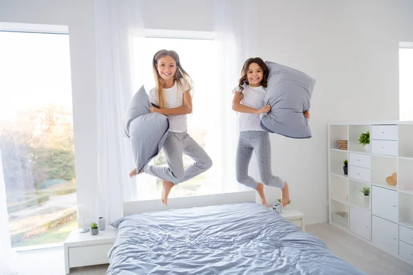 Πλήρες μέγεθος φωτογραφία των αστεία παιδιά κρατούν μαξιλάρια χέρι άλμα κρεβάτι δωμάτιο σε εσωτερικούς χώρους — Φωτογραφία Αρχείου