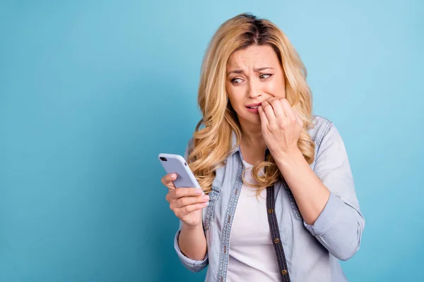 Retrato de la bella dama decepcionada mordiéndose los dedos mirando la pantalla del dispositivo aislada sobre fondo azul —  Fotos de Stock