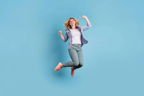 Full size photo of cute pretty woman jumping raising fists wearing pants trousers isolated over blue background — Stock Photo, Image
