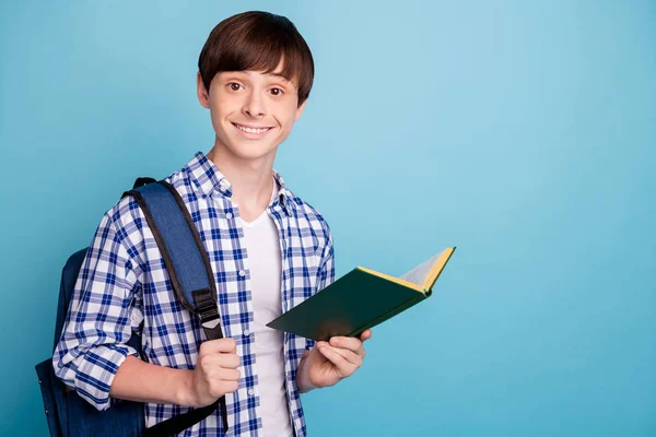 Портрет гарненького хлопчика, який тримає друковану книгу, усміхнену перевірену сорочку, ізольовану на синьому фоні — стокове фото