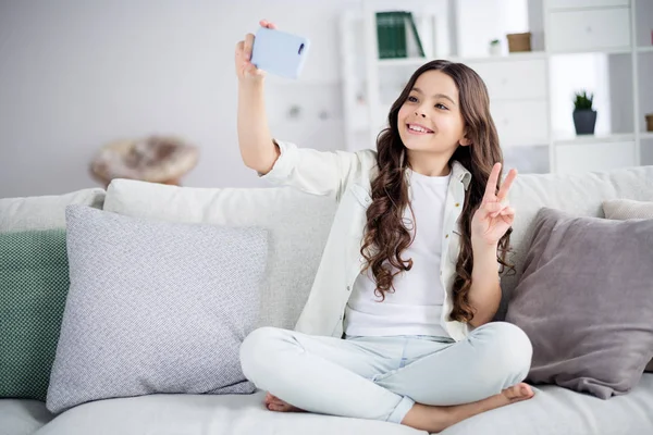 Portrait d'elle elle agréable attrayant charmant mignon joyeux gai positif ondulé cheveux fille preteen assis sur divan prendre faire selfie montrant v-signe dans la lumière blanche salon intérieur intérieur — Photo