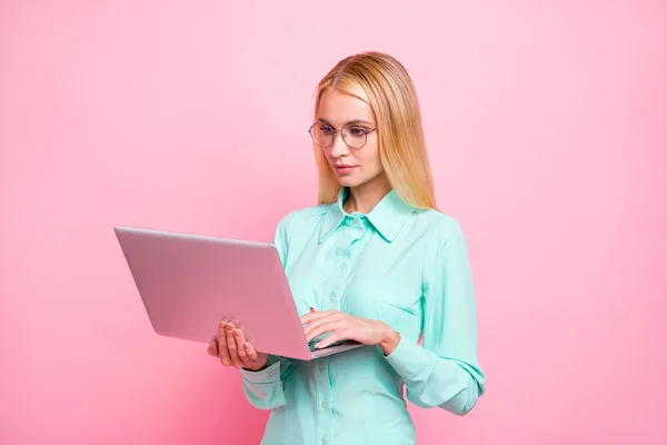 Портрет сфокусированной дамы в очках с современными технологиями, смотрящей на экран в бирюзовой рубашке, изолированной на розовом фоне — стоковое фото