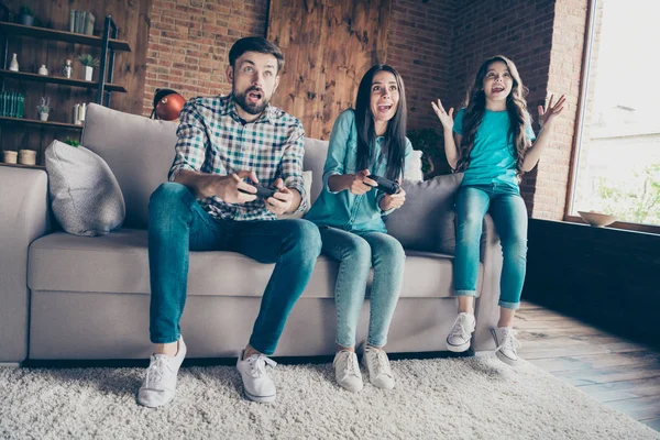 惊讶的已婚人士玩视频游戏和小孩坐在divan穿着牛仔牛仔裤格子衬衫在室内的房子的肖像 — 图库照片