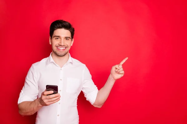 Foto do homem morena apontando para algo perto dele segurando o telefone com a mão enquanto isolado com fundo vermelho — Fotografia de Stock