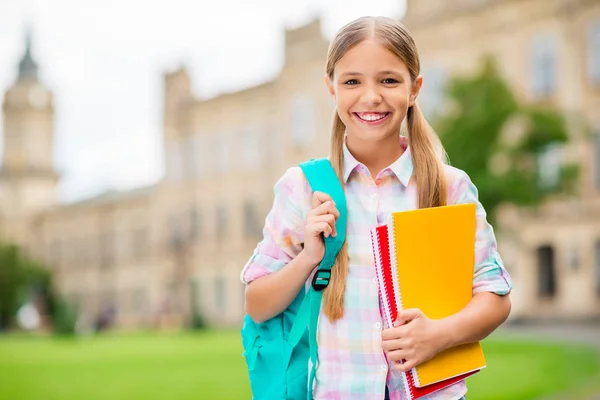Portrait of charming child holding books smiling wearing checkered plaid t-shirt standing outdoors — Stock Photo, Image