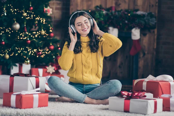Фотографія повного розміру тіла радісної приємної насолодженої дівчини, що слухає різдвяну музику, радіє новорічній атмосфері в жовтому джинсі з пуловеру, що сидить на підлозі — стокове фото