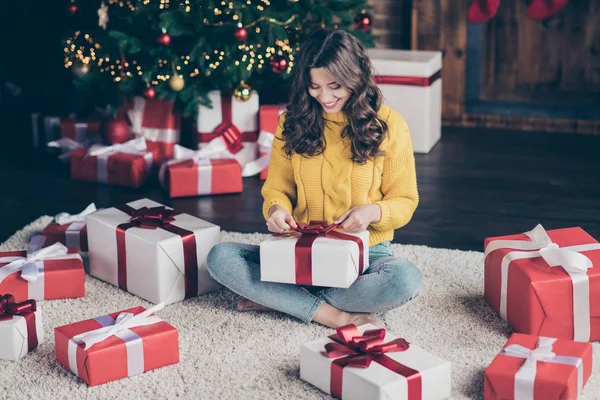 Fotka rozkošné milé dívky, která nosí džíny a žlutý svetr si vybalila svůj nový rok od Santa Clause, zatímco sedí na koberci na podlaze s nožičky zkřížené — Stock fotografie