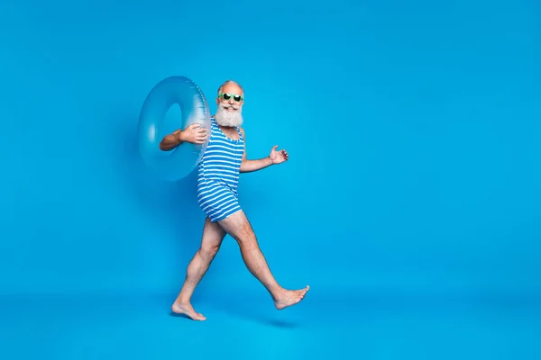 Foto de tamaño completo del tamaño del perfil del hombre alegre con gafas gafas que sostiene el anillo de juguete en su hombro caminando usando ropa de baño aislada sobre fondo azul — Foto de Stock