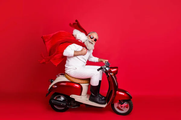Foto lateral de perfil de longitud completa del divertido pensionista que lleva el bolso con regalos sentado en la motocicleta que se apresura a usar botas de pantalones de suéter blanco aisladas sobre fondo rojo —  Fotos de Stock