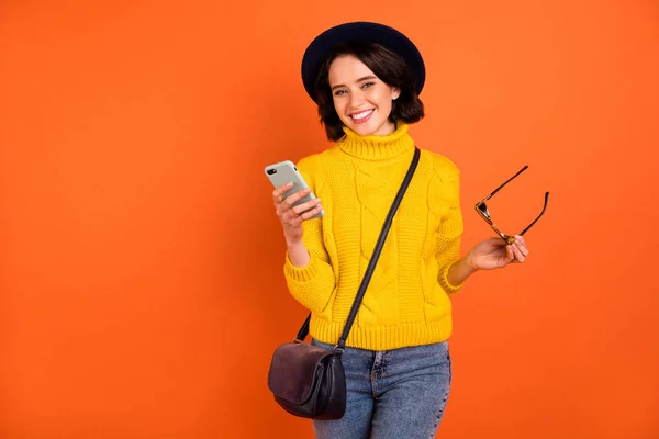 Foto van vrolijke mooie getande glimlachend meisje poseren in de voorkant van de camera met de telefoon in de hand terwijl geïsoleerd met oranje achtergrond — Stockfoto