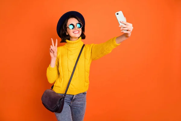 Foto di fotografare ragazza che indossa berretto berretto blogging con il suo nuovo telefono in maglione giallo che mostra simbolo v-segno isolato con sfondo arancione — Foto Stock