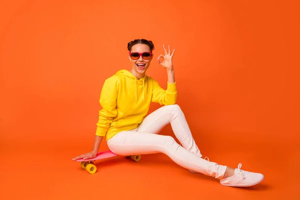 Retrato de jovens alegres mostrando sinal ok sentado no skate em calças brancas óculos de camisola amarela óculos isolados sobre fundo laranja — Fotografia de Stock