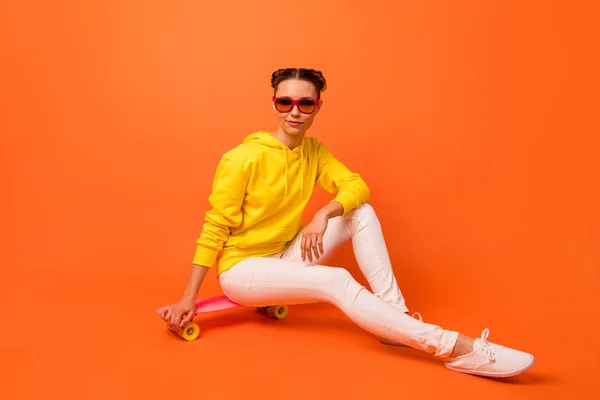 Retrato de chic millennial mirando sentado en skateboard desgaste pantalones jersey gafas gafas aisladas sobre fondo naranja — Foto de Stock