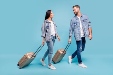 Full size profile side photo of two sweethearts carrying baggage seeing off wearing denim jeans jackets isolated over blue background clipart
