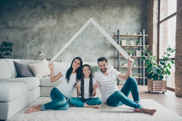 Portrait of nice attractive cheerful family in casual white t-shirts jeans sitting on carpet floor holding in hand roof real estate ownership showing v-sign at industrial loft style interior living — Φωτογραφία Αρχείου