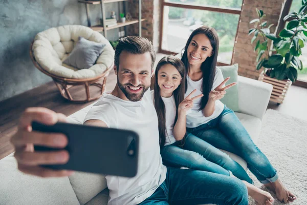 Mooie aantrekkelijke vrolijke vrolijke familie Mama Daddy dragen casual witte t-shirts jeans denim zittend op sofa maken selfie toont v-teken op industriële loft stijl interieur woonkamer — Stockfoto