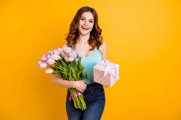 Retrato de senhora bonita segurando grande monte de tulipas rosa nos braços e giftbox desgaste tanque-top e jeans isolado fundo amarelo — Fotografia de Stock