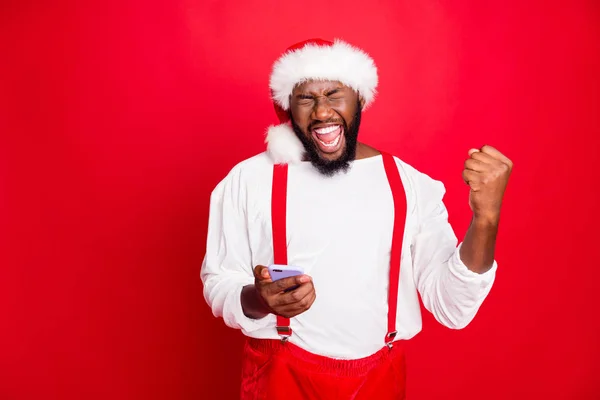 Porträt des verrückten afrikanischen Weihnachtsvaters, der die Fäuste hebt und schreit Ja bekam noble Nachrichten wie das Halten des Handys mit Hut, weißer Pullover isoliert über rotem Hintergrund — Stockfoto