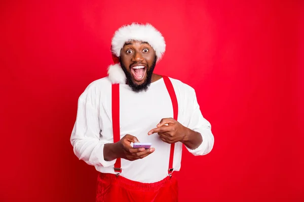 Retrato de Papai Noel preto impressionado em chapéu xmas apontando para seu dispositivo de telefone celular gritando wow omg ficou como vestindo pulôver branco isolado sobre fundo vermelho — Fotografia de Stock