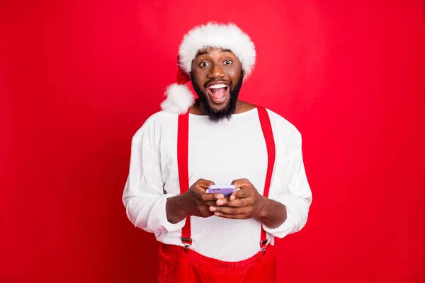 Porträt des beeindruckten schwarzen Weihnachtsmannes in Weihnachtsmütze schreiend wow omg Verwendung Handy moderne Technologie bekam noel Glückwünsche tragen weißen Pullover isoliert über rotem Hintergrund — Stockfoto