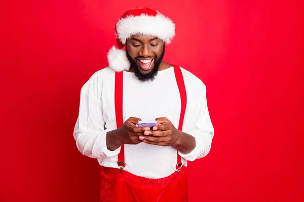 Retrato de la excitada raza mixta anta claus usando dispositivo de teléfono celular disfrutando de mensajes de año nuevo con gorra suéter blanco aislado sobre fondo rojo — Foto de Stock