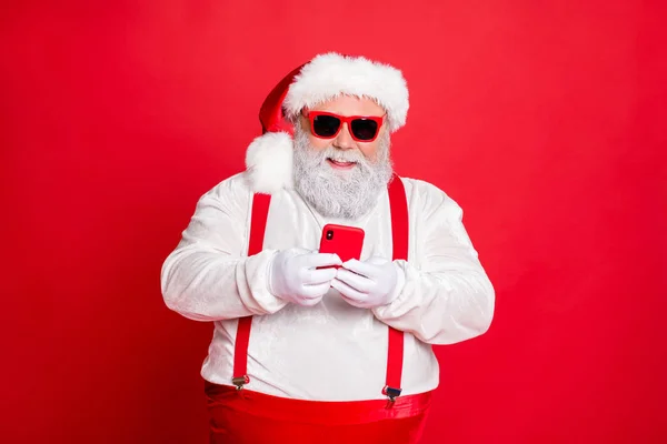 Ritratto di divertente grasso funky Babbo Natale in sovrappeso con grande addome pancia uso cellulare commento newyear post indossando occhiali alla moda occhiali cappello lettura notizie isolato su sfondo rosso — Foto Stock