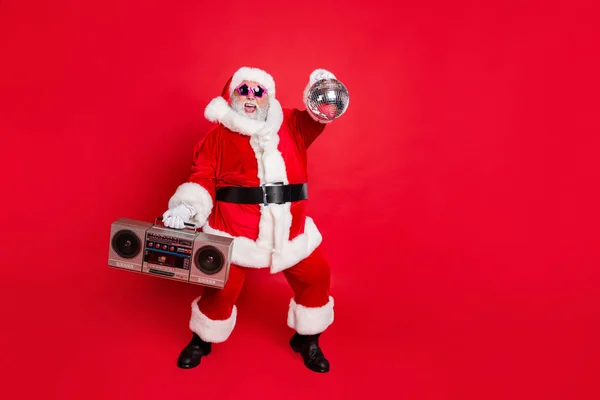 Foto de comprimento total de alegre Papai Noel em chapéus de cabeça segurando caixa de boom brilhante bola dançando na festa do ano novo vestindo estilo óculos de óculos elegantes isolados sobre fundo vermelho — Fotografia de Stock