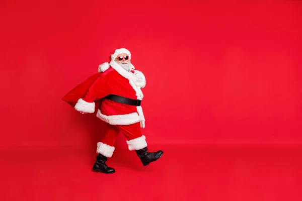 全长照片的梦幻般的脂肪有趣的圣诞老人有眼镜去携带袋与礼物在午夜隔离在红色背景 — 图库照片