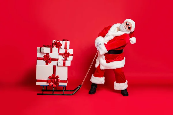 Fulll foto de longitud de Santa Claus cansado tirando de su trineo con paquetes de regalos en la medianoche con gafas de gafas cinturón de gafas aislado sobre fondo rojo — Foto de Stock