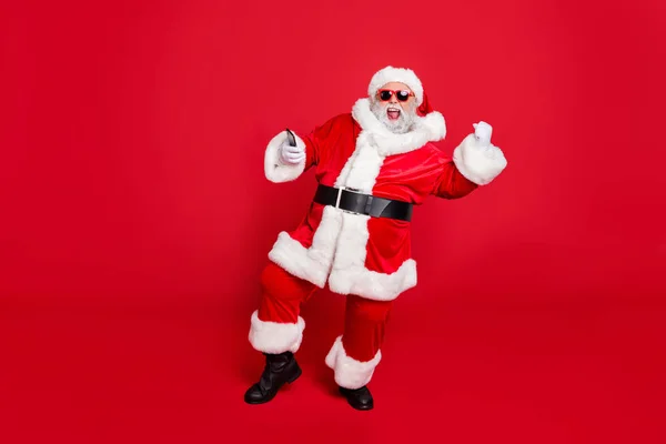 Per sempre giovane. Foto a figura intera di Babbo Natale eccitato in cappello utilizzando il dispositivo cellulare alzando i pugni urlando sì hanno partito indossando occhiali da cintura occhiali isolati su sfondo rosso — Foto Stock