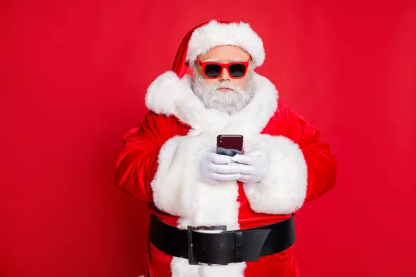 Portrait de Père Noël esprit claus avec des lunettes de vue à l'aide de téléphone cellulaire dispositif de lecture nouvelles portant costume isolé sur fond rouge — Photo