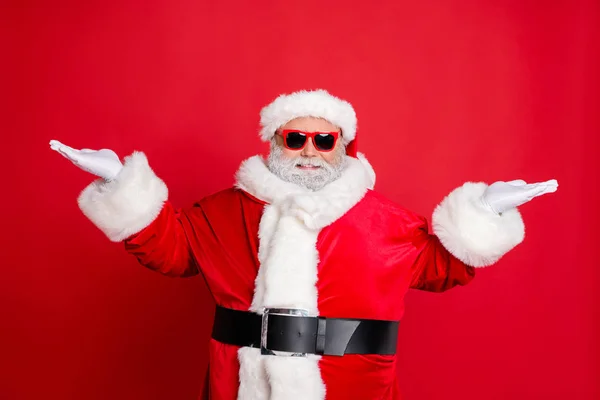 Retrato de bonito legal engraçado santa claus com óculos segurando anúncios do ano novo promo vestindo tampa do cinto preto isolado sobre fundo vermelho — Fotografia de Stock