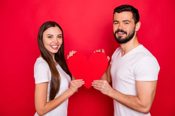 Foto de encantadora linda pareja encantadora de dos personas juntas sosteniendo el corazón con sus manos usando una camiseta blanca mientras están aisladas con fondo rojo — Foto de Stock