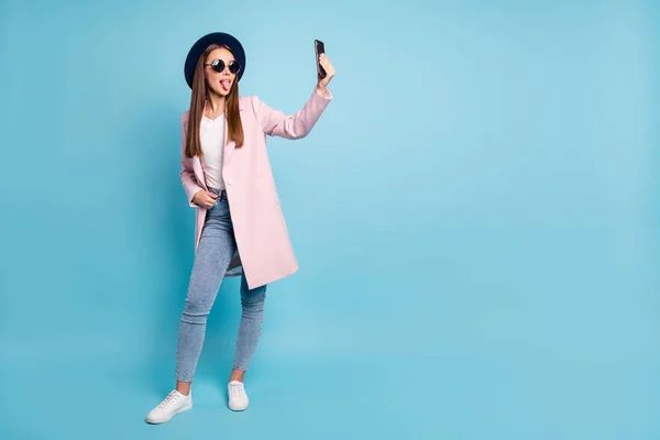 Foto in voller Größe von kindischen Mädchen mit ihrem Handy machen Selbstfoto Spaß mit Brille Brille Topcoat Jeans isoliert über blauem Hintergrund — Stockfoto