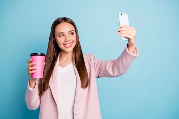 Портрет милой веселой леди с помощью мобильного телефона делает селфи держа кружку с горячим напитком носить розовое пальто изолированы на голубом фоне цвета — стоковое фото