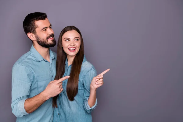 Retrato de dos encantadores hombres y mujeres casados señalando el espacio de copia después de la promoción con jeans camisa aislada sobre fondo gris —  Fotos de Stock