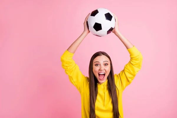 Foto de encantador bonito namorada alegre segurando bola de futebol acima de sua cabeça vestindo suéter amarelo, enquanto isolado sobre fundo cor pastel rosa — Fotografia de Stock