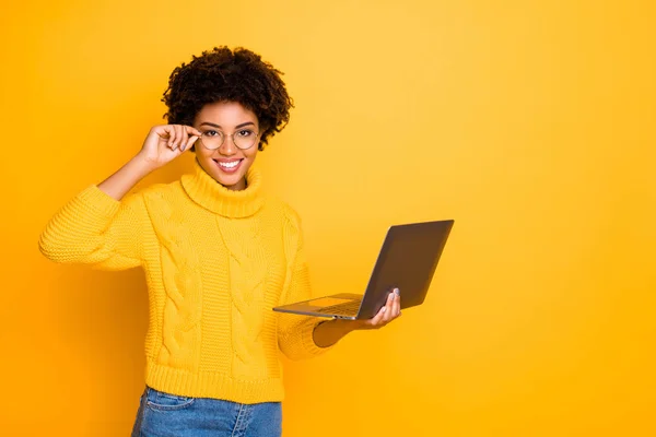 Photo de charmant mignon noir bouclé jolie petite amie tenant son ordinateur portable et lunettes à la recherche de quelque chose obtenir aime porter un jean pull jaune isolé couleur vive arrière-plan — Photo