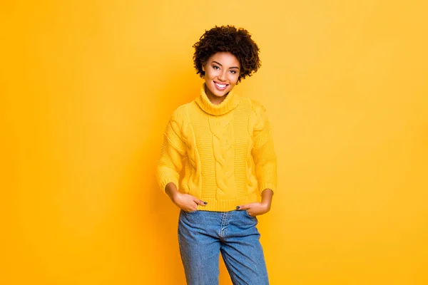 Foto de señora divertida de piel oscura en buen humor desgaste cálido jersey de punto y pantalones vaqueros de pie aislado de color amarillo fondo — Foto de Stock