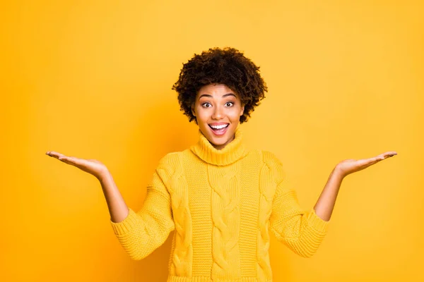 Foto de la señora de las ventas de piel oscura sosteniendo dos nuevos productos en los brazos proponiendo los mejores precios usar el suéter de punto caliente aislado fondo amarillo — Foto de Stock