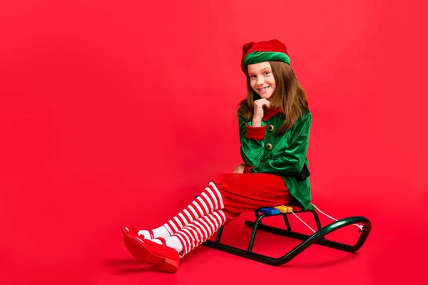 Porträtt av henne hon trevlig attraktiv charmig glad glada roliga söta pre-Teen Elf jultomten kläder ser outfit sitter på släde isolerad över ljusa levande Shine röd bakgrund — Stockfoto
