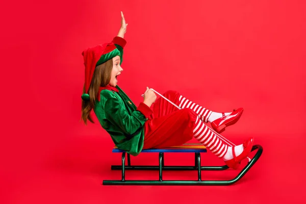 Perfil de corpo inteiro foto lateral da menina elfo louco com cabeça vermelha longo corte de cabelo trenó se divertindo vestindo chapéu de traje verde chapéu isolado sobre fundo vermelho — Fotografia de Stock