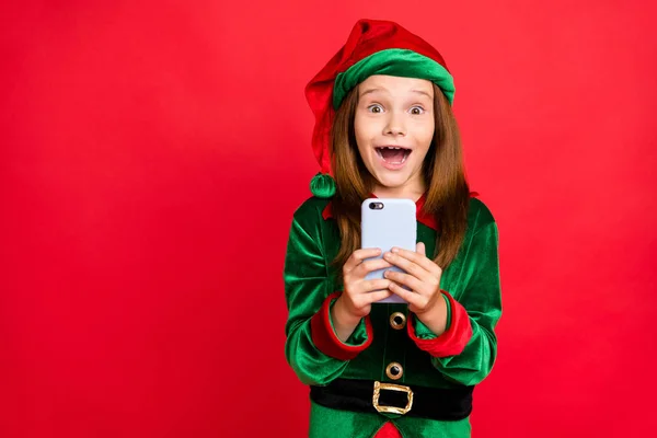 Porträtt av chockad Elf i grön hatt mössa med långa röda huvudet skrikande wow OMG använda smartphone få som Noel kommentar bär Elf kostym isolerad över röd bakgrund — Stockfoto