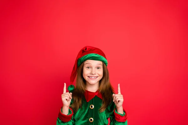 Portrait of cheerful elf in green cap smiling showing ads with her index finger recommend party wearing fairy costume isolated over red background — Zdjęcie stockowe