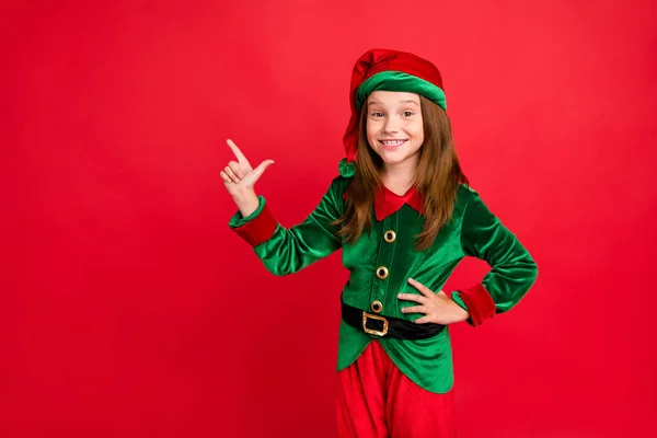 Portrait of lovely child elf in hat showing her finger point at ads recommend party wearing green elf costume hat cap isolated over red background — Stockfoto