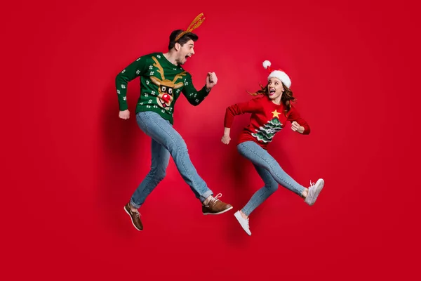 Full body photo of excited jumping couple rushing for x-mas discounts wear ugly ornament jumpers isolated red color background — Stockfoto