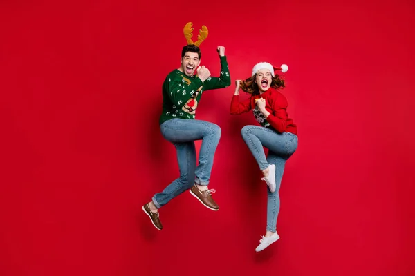 Foto de cuerpo completo de la pareja de salto sorprendido excitado por los precios de x-mas usan jerséis adornos feos y sombreros aislados de color rojo de fondo —  Fotos de Stock