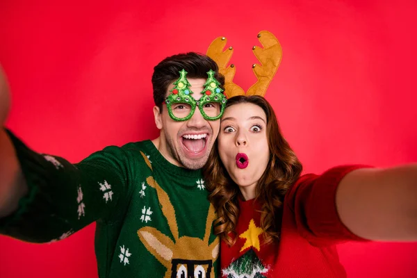 Photo of excited lady and guy chilling at x-mas costume party making selfies wear funky pullovers with ugly ornaments isolated red color background