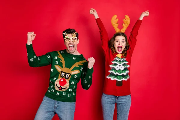 Retrato de loco divertido dos esposa de la familia marido pelo castaño celebrar la victoria levantar los puños gritar sí usar árbol de navidad diseño jersey jeans traje gafas aislado sobre fondo de color rojo —  Fotos de Stock