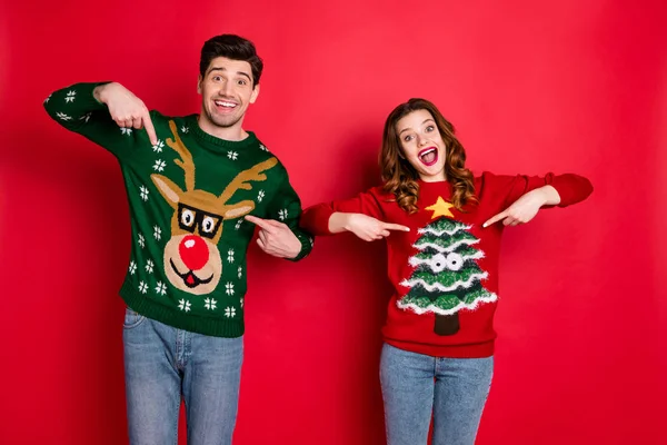 Porträt der erstaunten Familie zwei Personen brünettes welliges Haar Ehefrau Ehemann zeigt Zeigefinger seinen gemütlichen Pullover mit Hirsch Weihnachtsbaum Design Jeans isoliert über rotem Hintergrund — Stockfoto